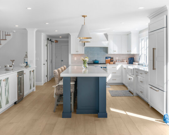 Sunlit wood floor in kitchen with white cabinets and blue island with dining chairs
