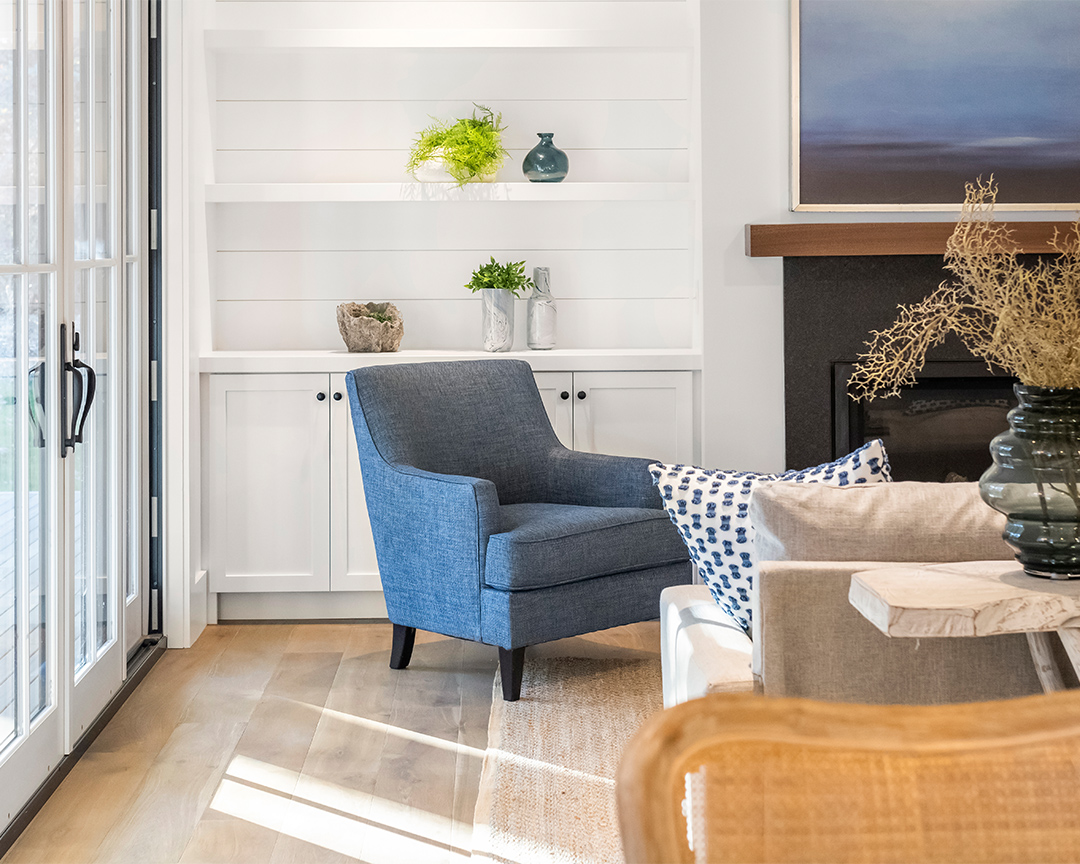 A cozy living room space with coastal design elements, featuring white shiplap walls and built-in shelving units for displaying decorative items. A sunlit dark brown wood floor completes the space.