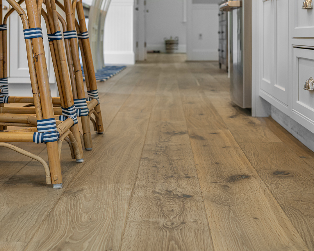 Close up of kitchen floor with wide planks, showing the bottom of white cabinets and rattan chairs