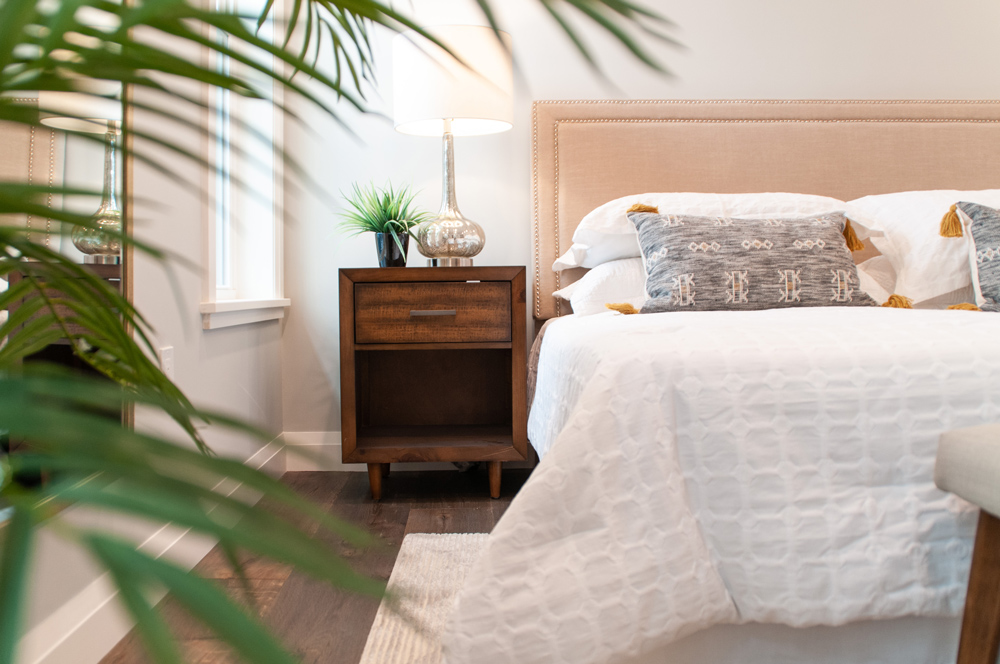 Close up of plant leaves with bed and end table in view, as well as a dark brown wood floor.