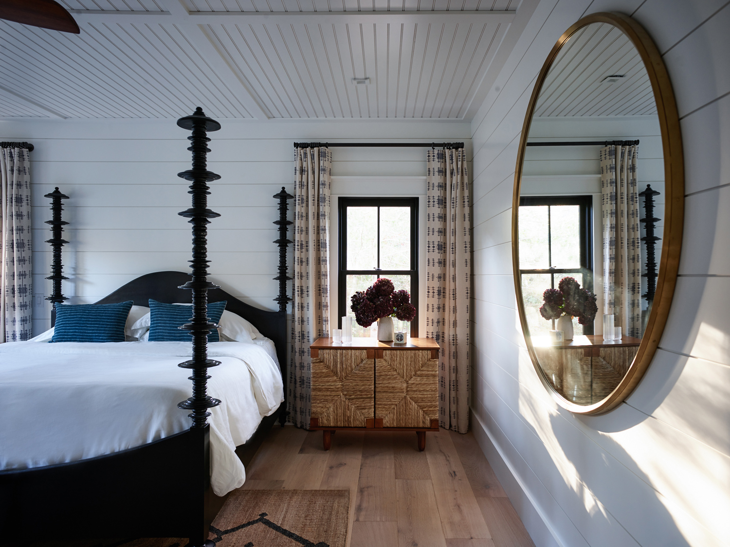 Cozy bedroom and bed with tall posts, round mirror, side table, and dark brown wood flooring.