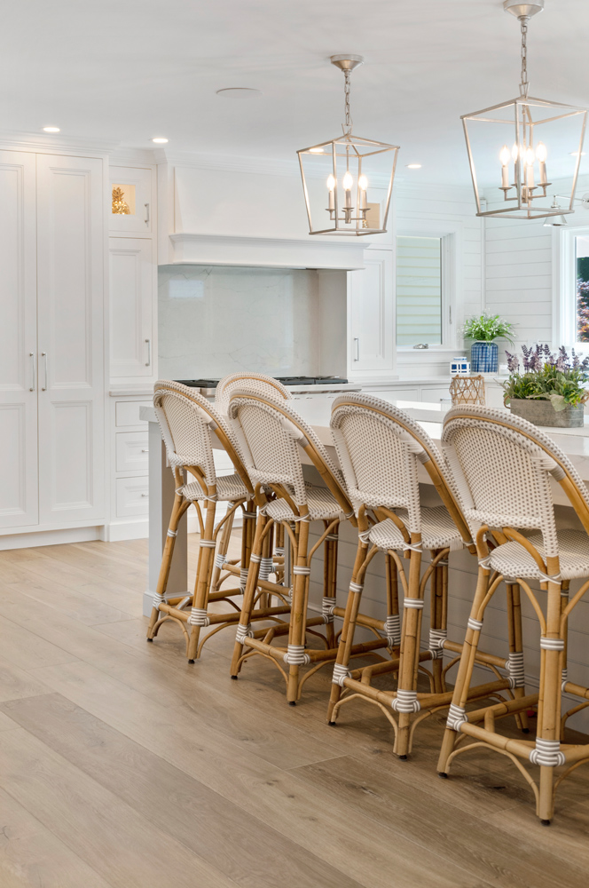 Rattan chairs in a white kitchen with pendant lights, decorative plants, and wide plank wood flooring.