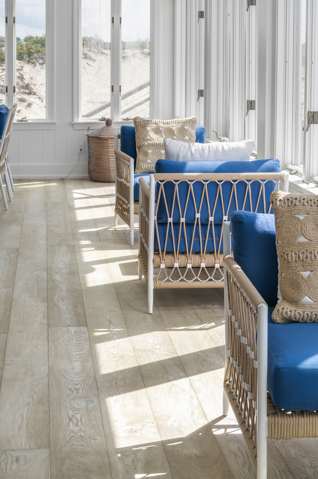 Sunlit wood floor in sitting area with blue chairs and beach visible from windows.